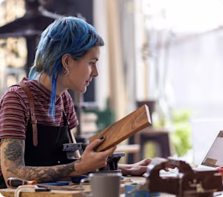 Tattooed person with blue hair holding a piece of wood while working on a laptop in a workshop.